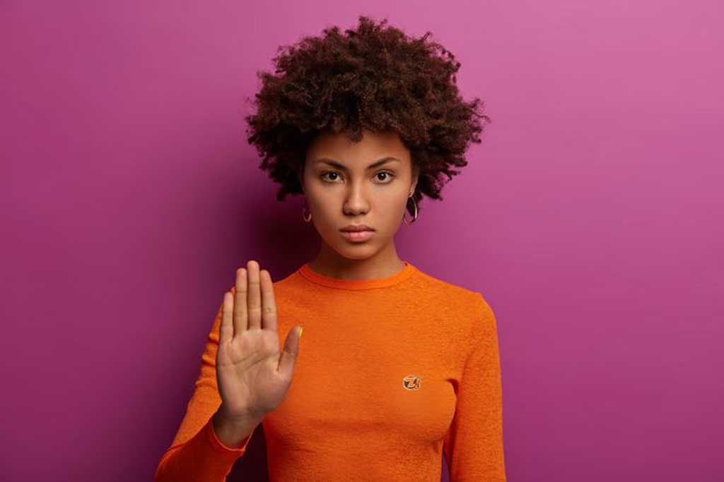 Mulher séria faz gesto de pare, veste suéter laranja isolado na parede roxa ilustra nosso artigo sobre: Qual a importância de estabelecer limites?